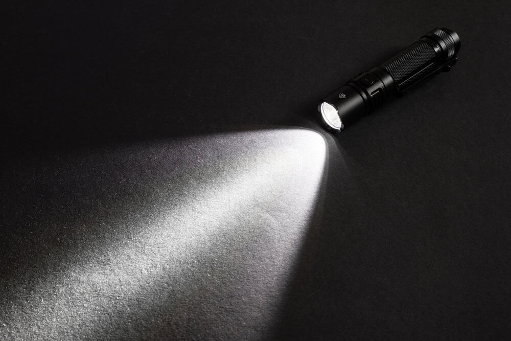 A powerful black tactical flashlight cuts through darkness with a focused white beam.