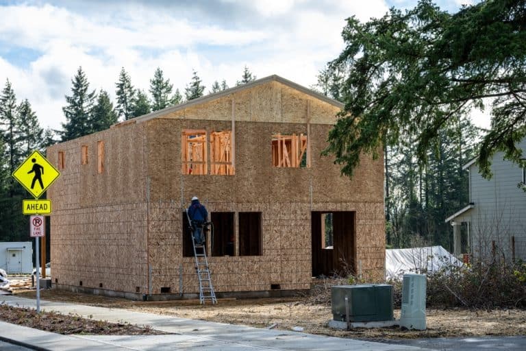 A worker climbs a ladder on a new house under construction in a serene, wooded neighborhood.