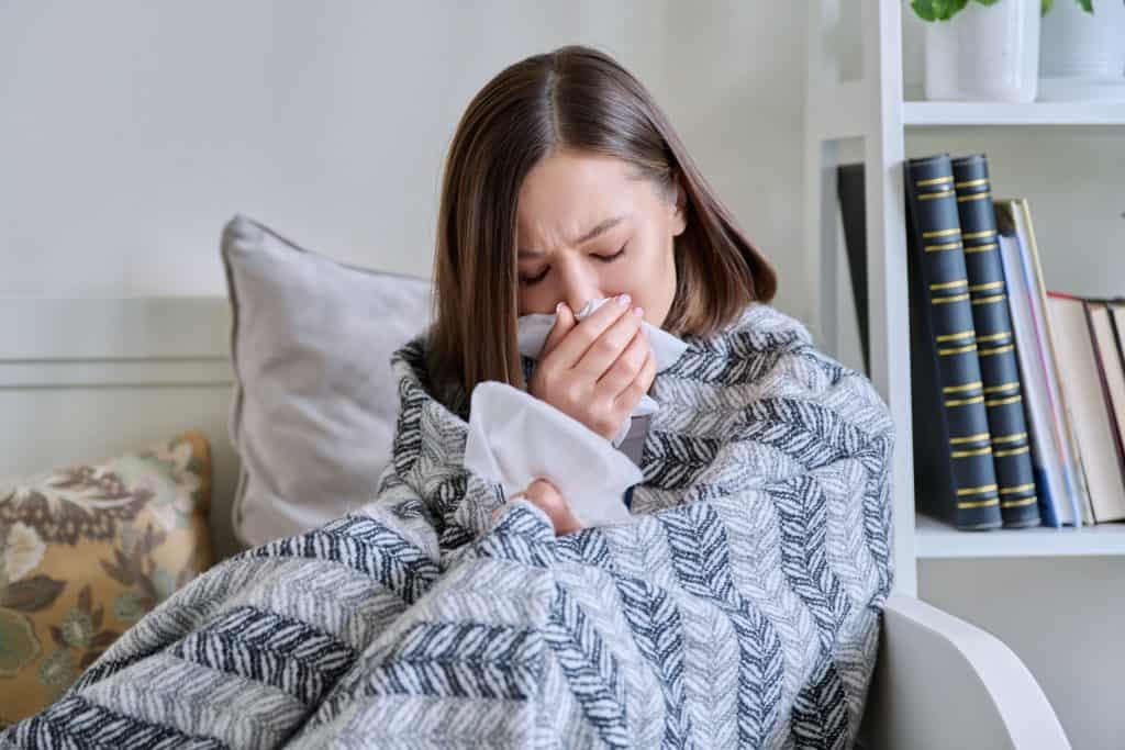 A woman wrapped in a patterned blanket sits on a couch, holding a tissue to her face while sneezing. The environment appears cozy, with pillows and bookshelves in the background, but her expression shows discomfort, possibly due to allergies or indoor air pollutants.