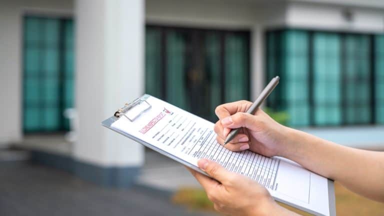 Home inspection in progress: hands fill out a property evaluation form on a clipboard outside a modern house.