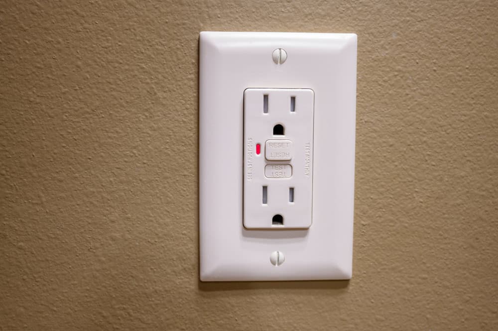 A GFCI outlet with a glowing red indicator light, signaling a tripped circuit on a tan wall.