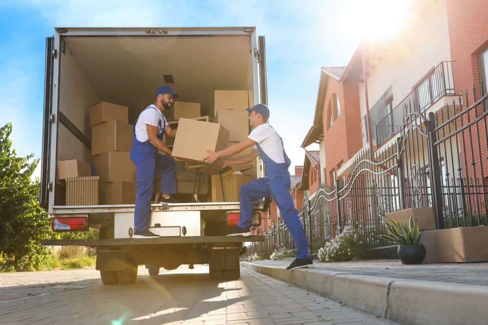 Two workers in blue uniforms loading boxes into a moving truck on a sunny street.