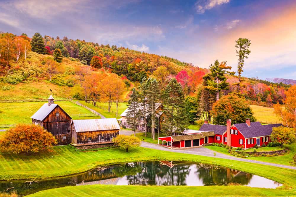 A serene autumn scene with vibrant foliage, rustic barns, and a charming red farmhouse reflected in a tranquil pond.