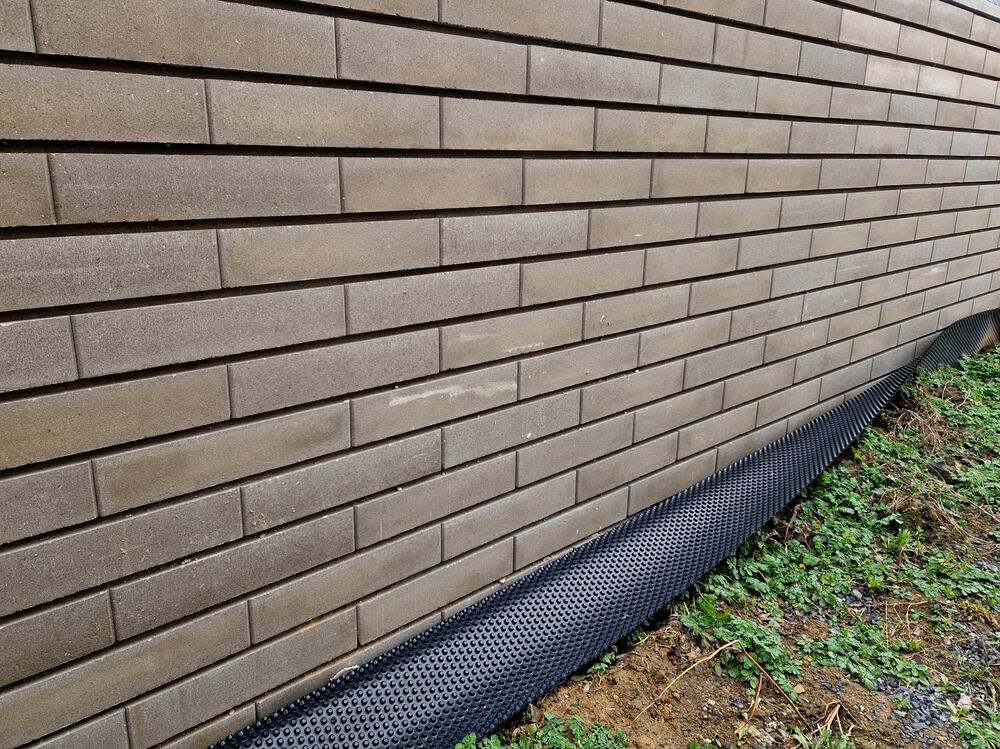 A modern brick wall with drainage mesh at its base, surrounded by greenery.