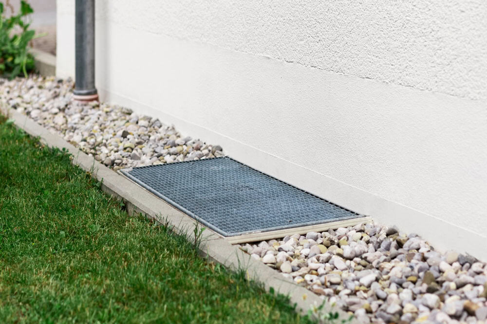 A drainage grate nestled among pebbles beside a white wall, bordered by a strip of green grass.