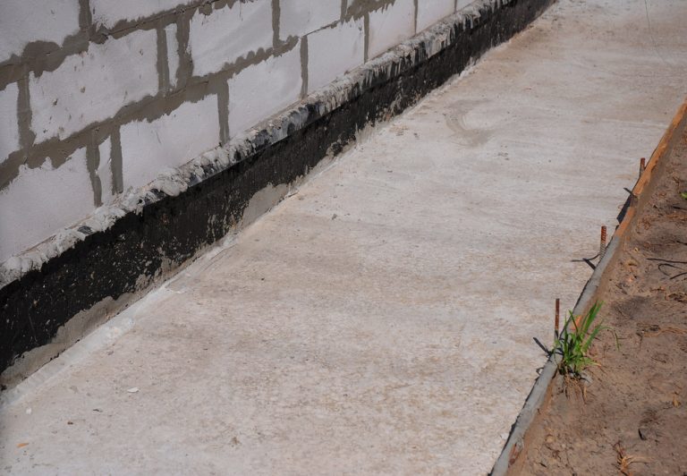 Freshly poured concrete walkway beside a cinder block foundation with black waterproofing.