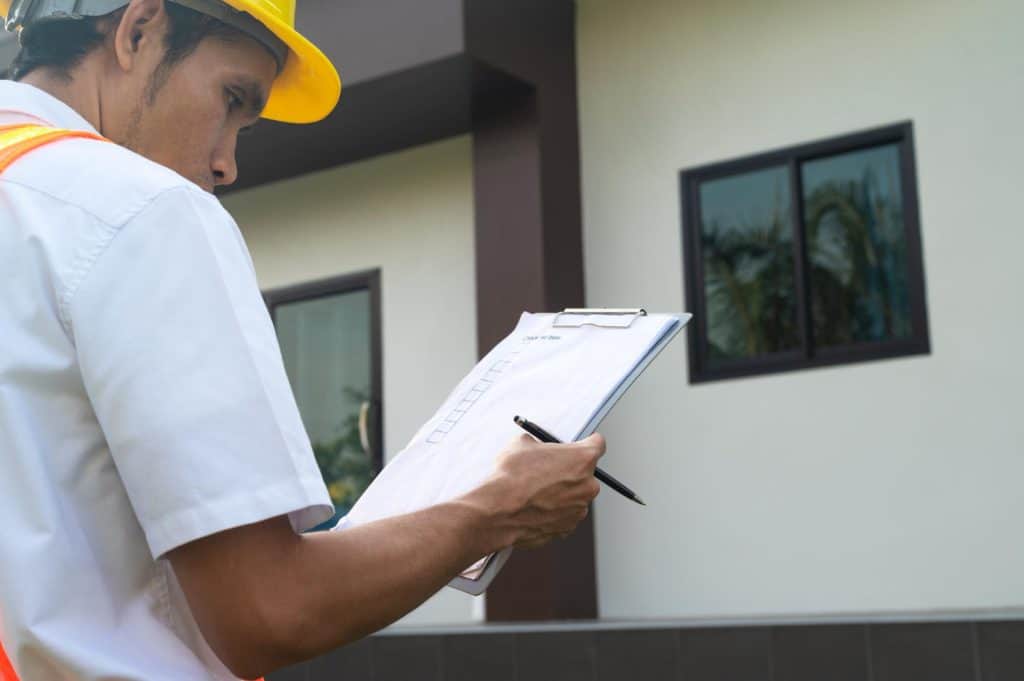 Construction inspector in a yellow hardhat reviews documents during a building site assessment, pen and clipboard in hand.