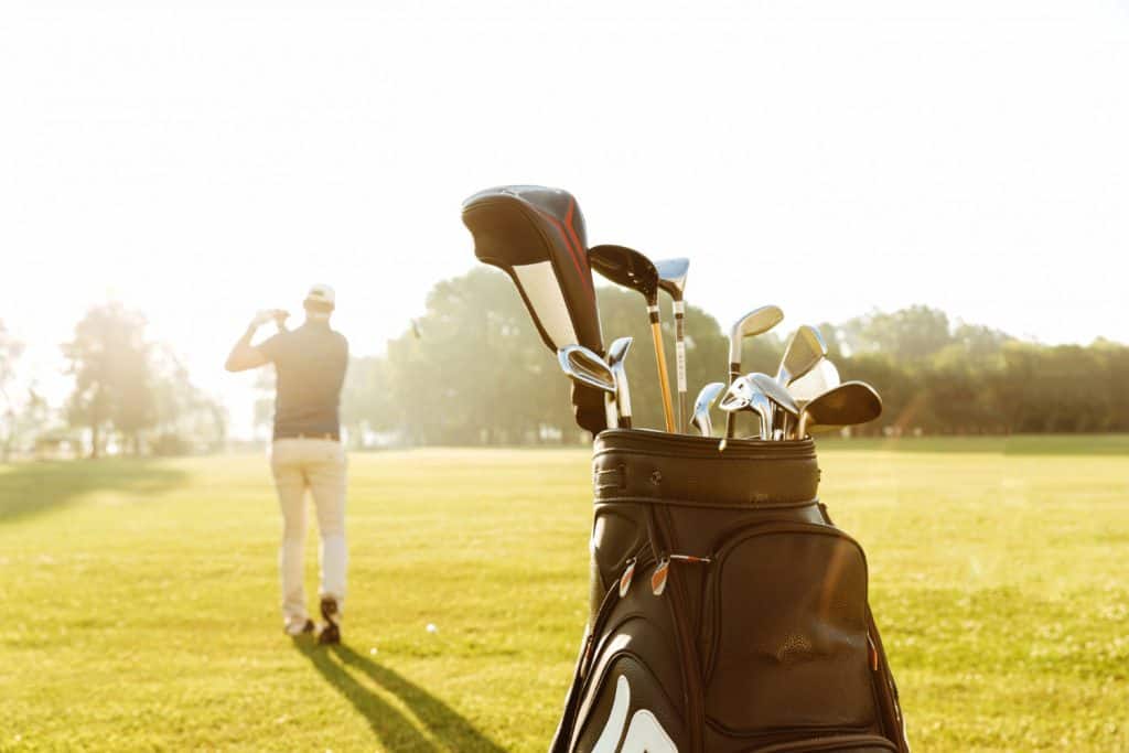 Golf bag filled with clubs rests on a sun-drenched course while a player takes their shot in the golden afternoon light.