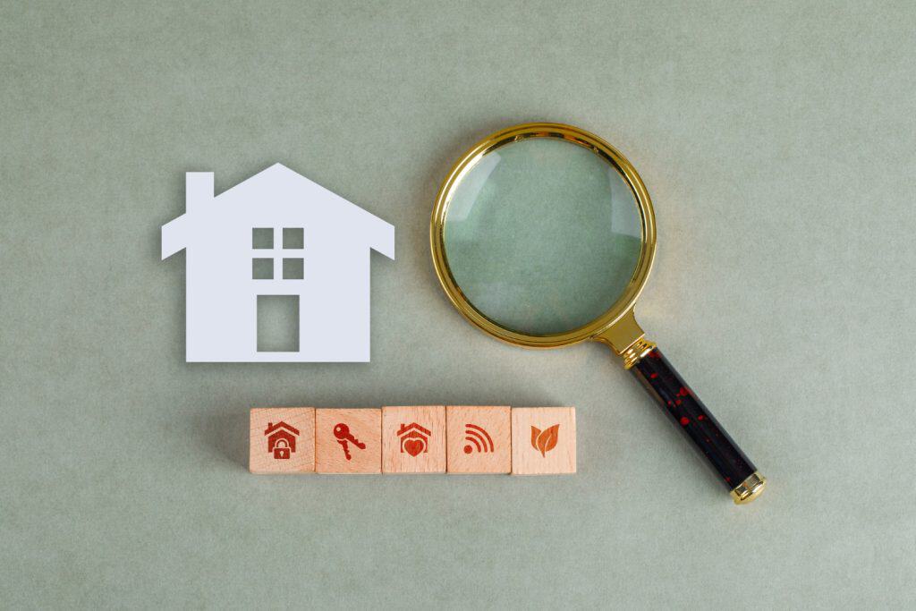 Home inspection essentials: paper house cutout, wooden blocks with property icons, and vintage magnifying glass on sage background.