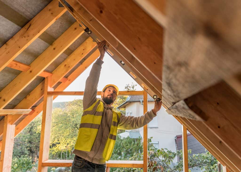 Construction worker in high-visibility vest measures wooden beams on a residential roof structure, examining the craftsmanship against the backdrop of a sunny suburban setting.