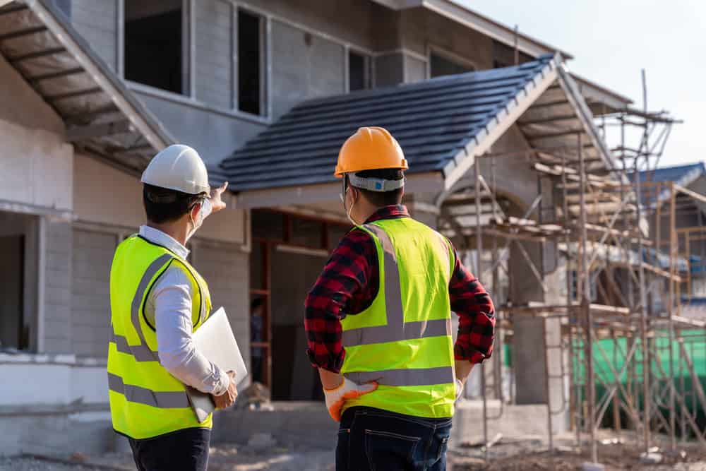Two construction workers in safety gear discuss plans in front of a house under construction.