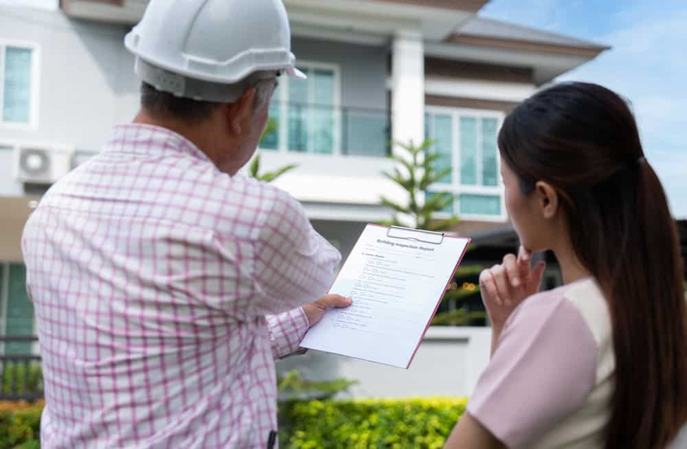 A contractor and a homeowner review a building inspection report in front of a modern house.