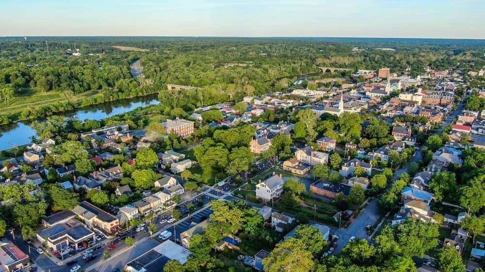 Historic Fredericksburg, Virginia from above, showing its charming downtown district nestled along the Rappahannock River, with its mix of colonial architecture, tree-lined streets, and historic stone railway bridge in the distance.
