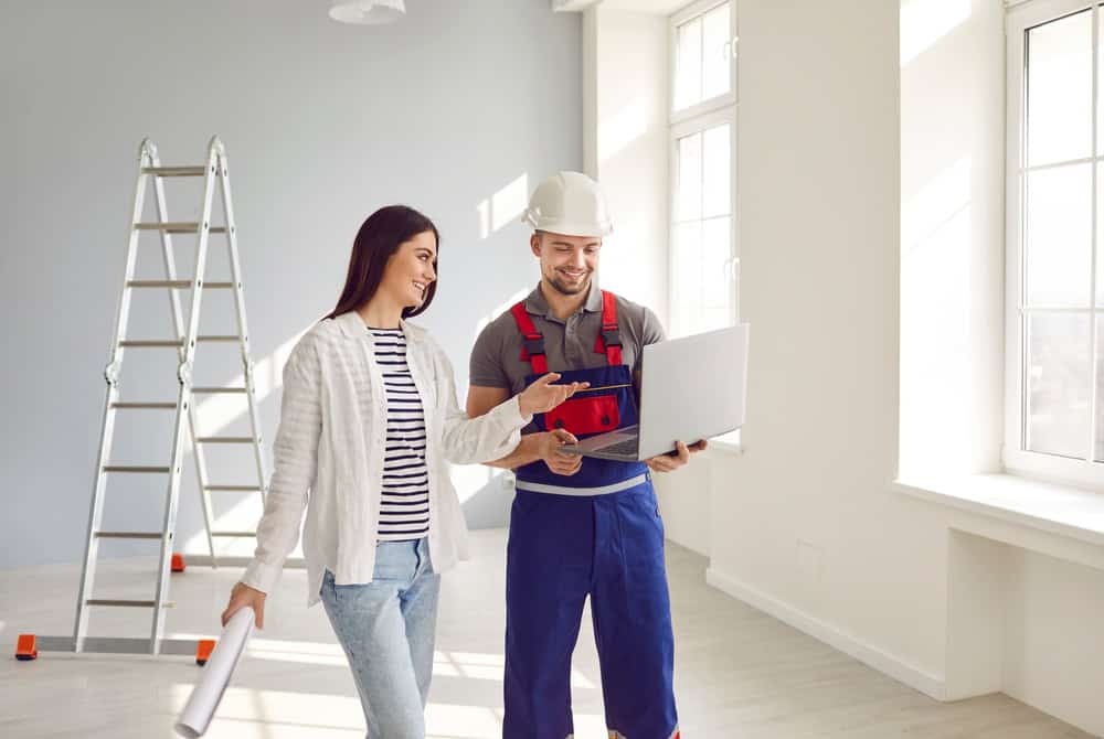 A woman and a construction worker collaborate on a project, reviewing plans on a laptop in a bright, newly constructed room.