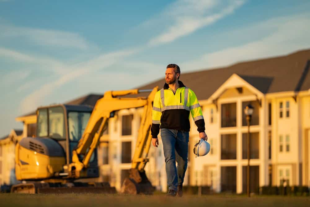 A construction worker walks confidently with a hard hat in hand, with a digger and residential buildings in the background.