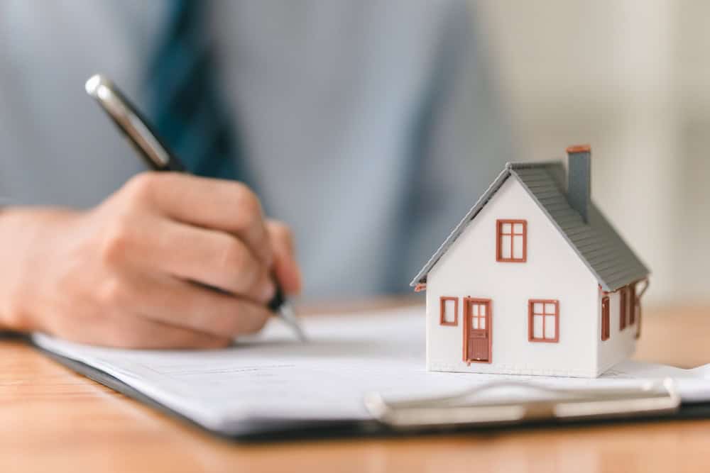 A small white model house with a gray roof and brown door and windows sits on a document, while a hand holds a pen, poised to sign what appears to be real estate paperwork. This scene captures the moment of finalizing a property transaction or mortgage agreement.