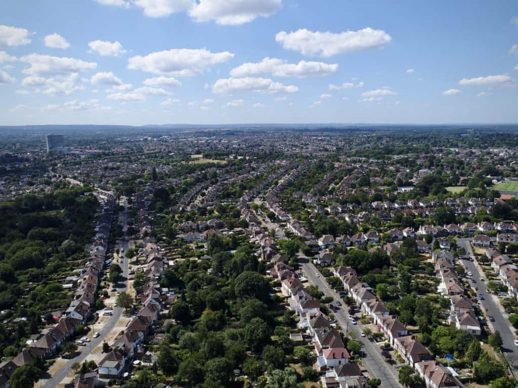 Aerial view of a suburban neighborhood with neat rows of semi-detached houses, tree-lined streets, and sprawling residential areas stretching toward the horizon under a cloud-dotted summer sky.