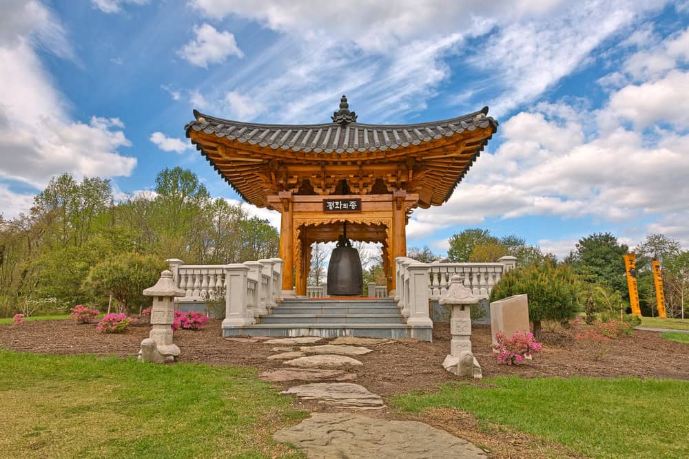 A traditional Korean pavilion with a large bell, surrounded by lush greenery and blooming flowers.