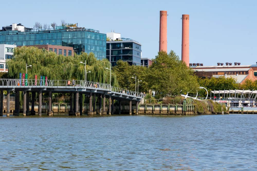 A scenic view of The Wharf in Washington, D.C., featuring the pedestrian bridge, lush greenery, and the iconic twin smokestacks in the background.