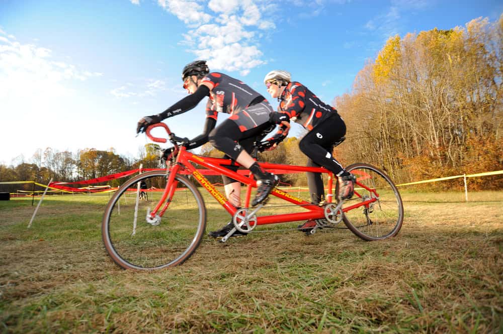 Two cyclists race on a tandem bike through a scenic autumn landscape.