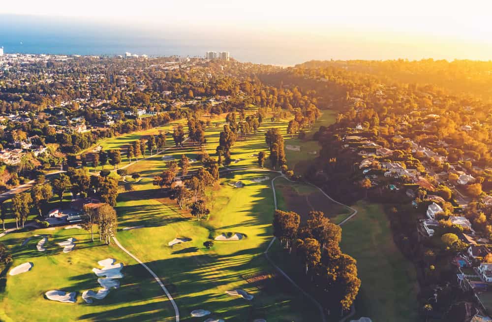 Aerial view of a lush golf course nestled among suburban homes with the ocean in the distance at sunset.