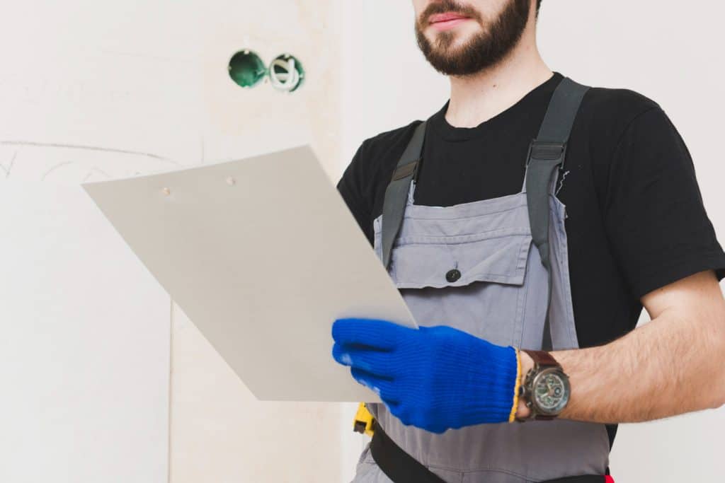 Construction professional in grey overalls and protective blue gloves reviewing documents on a clipboard during a site inspection.