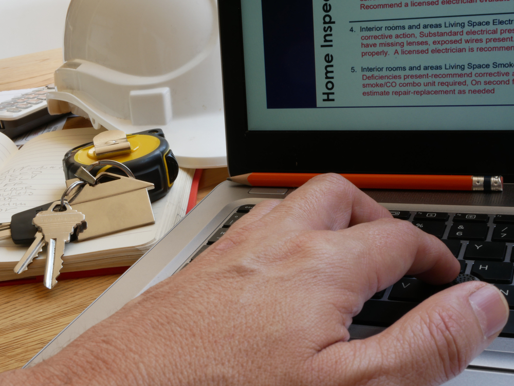 A hand types on a laptop, surrounded by a hard hat, keys, and a tape measure, indicating a home inspection report in progress.