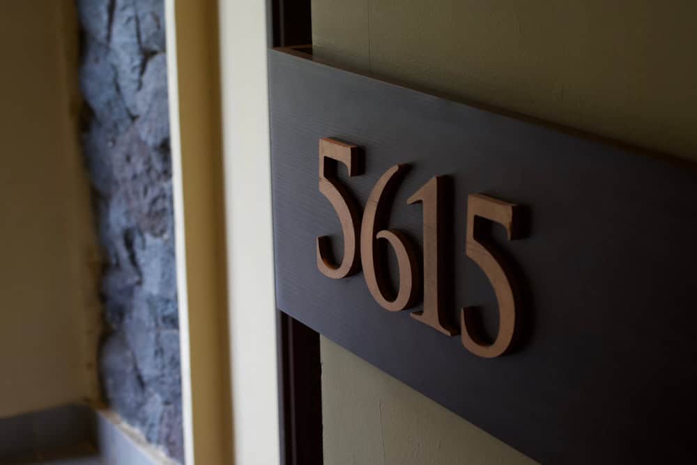 A stylish wooden room number "5615" mounted on a dark wood panel, likely for a hotel or apartment door. The elegant, dimensional numbers are set against a textured backdrop, with stone or textured wall visible to the left.
