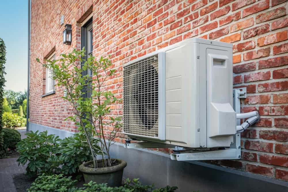 A modern air conditioning unit mounted on a rustic brick wall, surrounded by lush greenery and a quaint lantern.