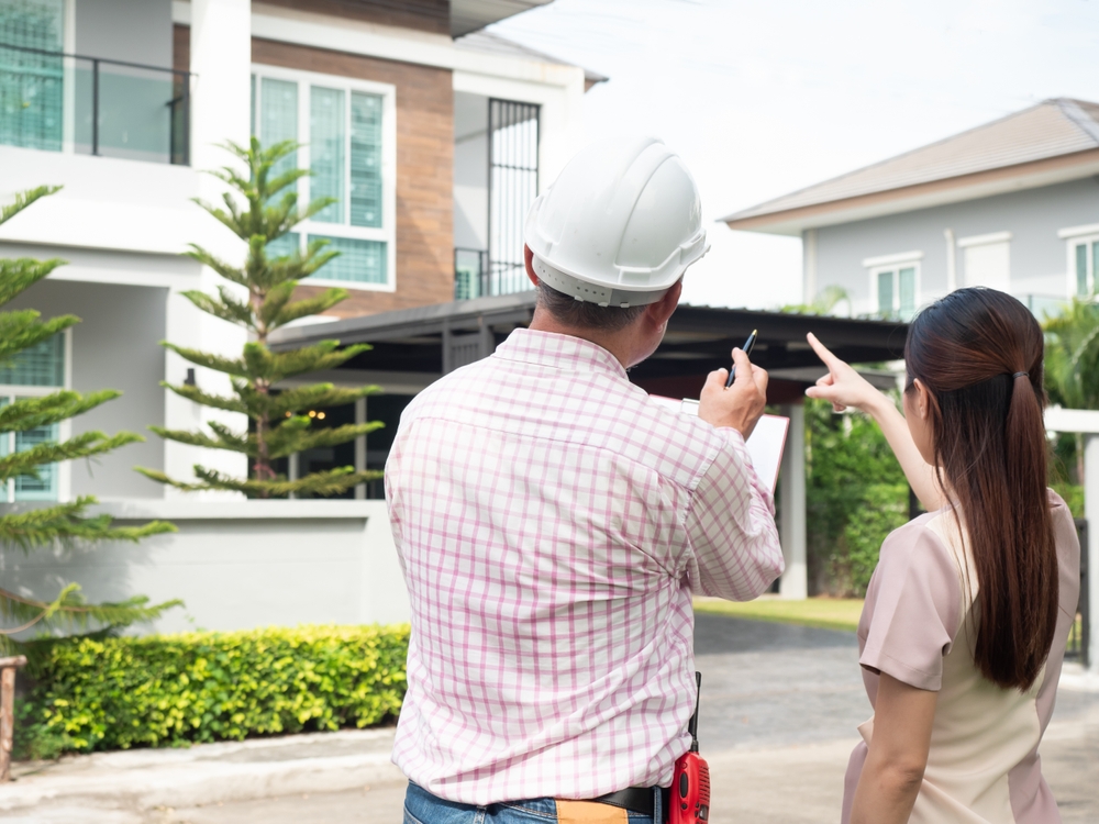 A construction worker and a woman discuss plans in front of a modern house, pointing and taking notes.