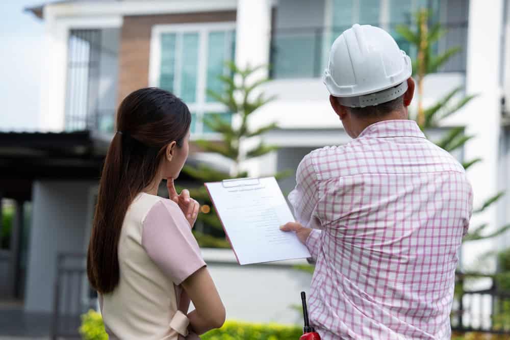 A home inspector and a client review blueprints in front of a modern house.