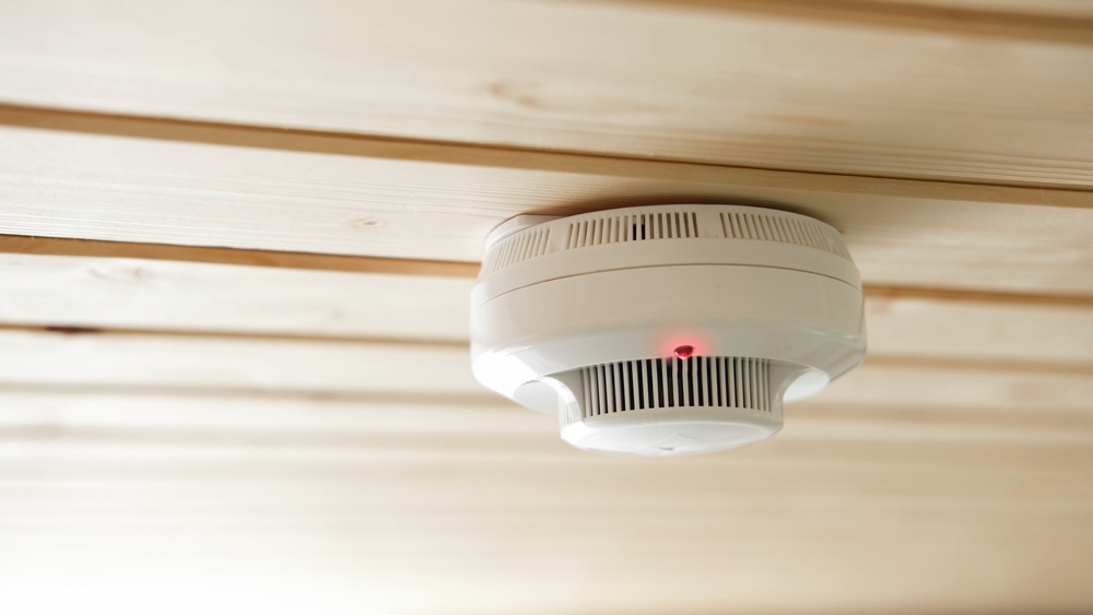 A smoke detector mounted on a wooden ceiling, with its red indicator light glowing.