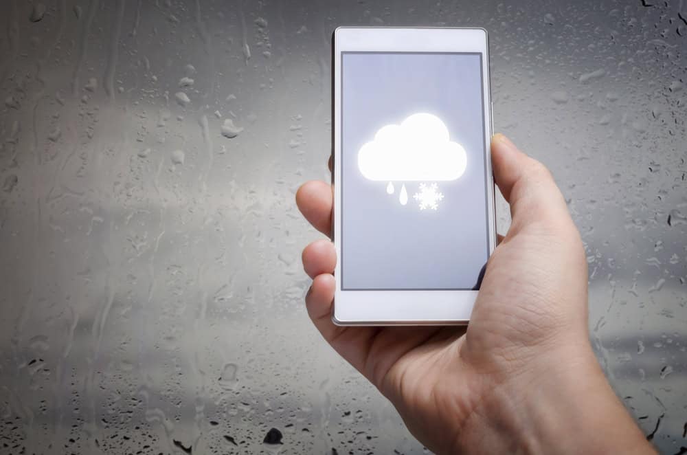 Hand holding a smartphone displaying a snowy weather forecast against a rainy window background.