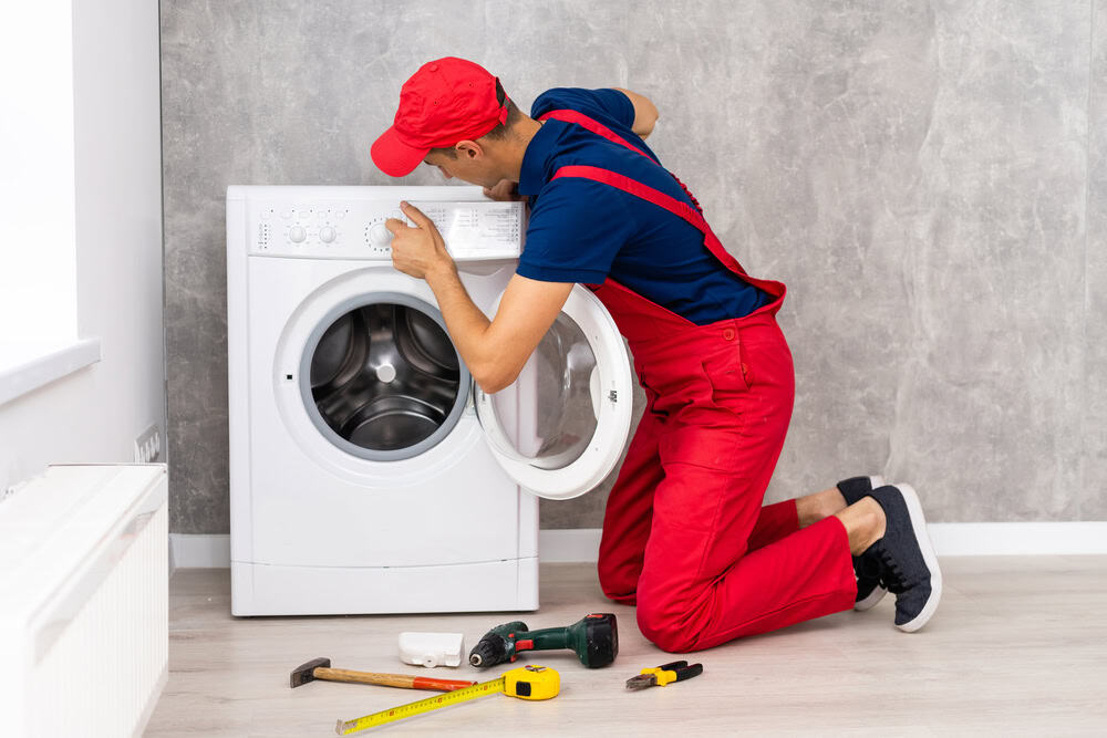 A technician in red overalls diligently repairs a washing machine, surrounded by tools on the floor.