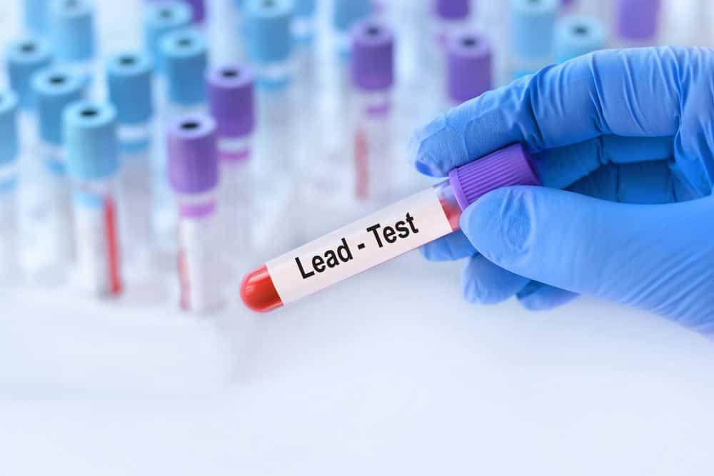A gloved hand holds a test tube labeled "Lead-Test" among other test tubes in a laboratory setting.