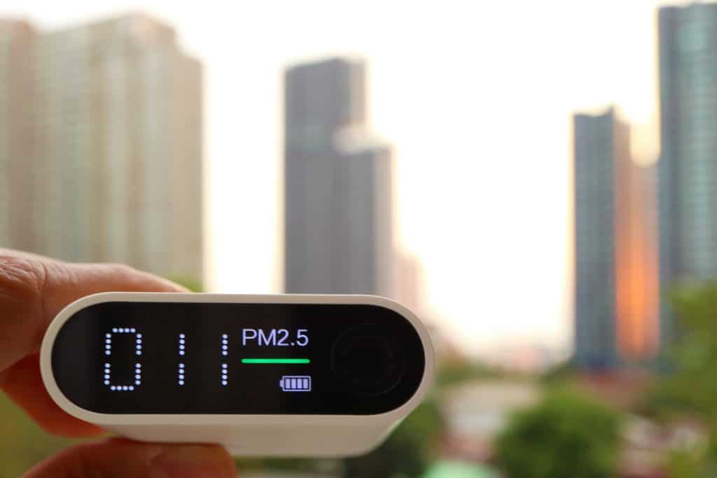 A hand holding a digital air quality monitor displaying PM2.5 levels with a cityscape in the background.