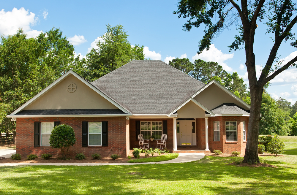 Charming brick ranch home nestled among lush greenery, featuring a welcoming front porch with rocking chairs and well-manicured lawn.