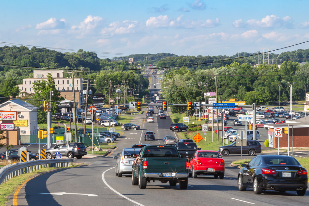 Busy suburban street scene with multiple cars driving on a curved road lined by powerlines and businesses on a sunny day.