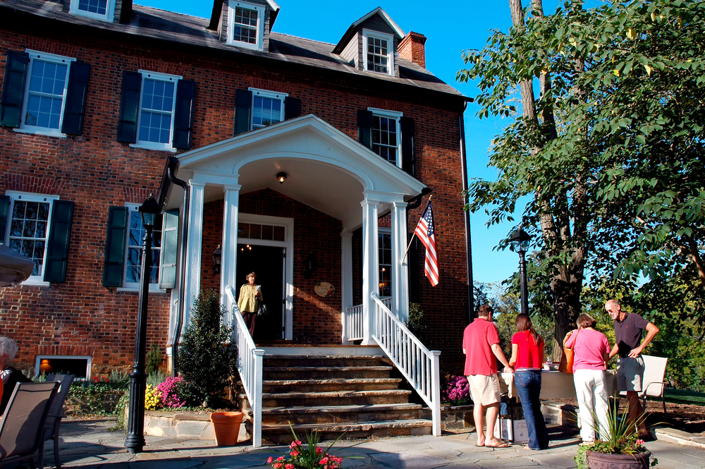 Visitors gather in front of a charming brick house adorned with an American flag and surrounded by lush greenery.