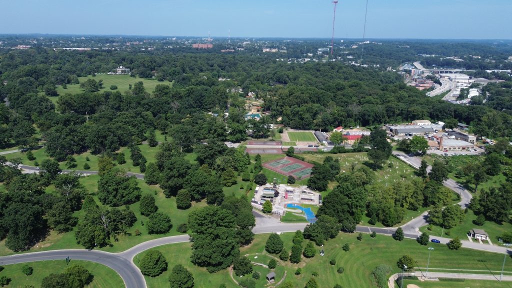 An aerial view of a sprawling park with tennis courts, a playground, and a cemetery, surrounded by lush greenery and a glimpse of a highway in the distance.
