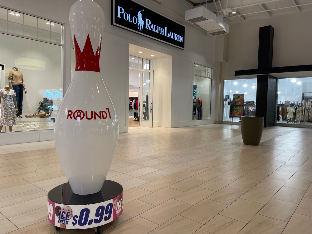 Giant bowling pin promoting $0.99 ice cream stands in a mall corridor, with stores like Polo Ralph Lauren in the background.
