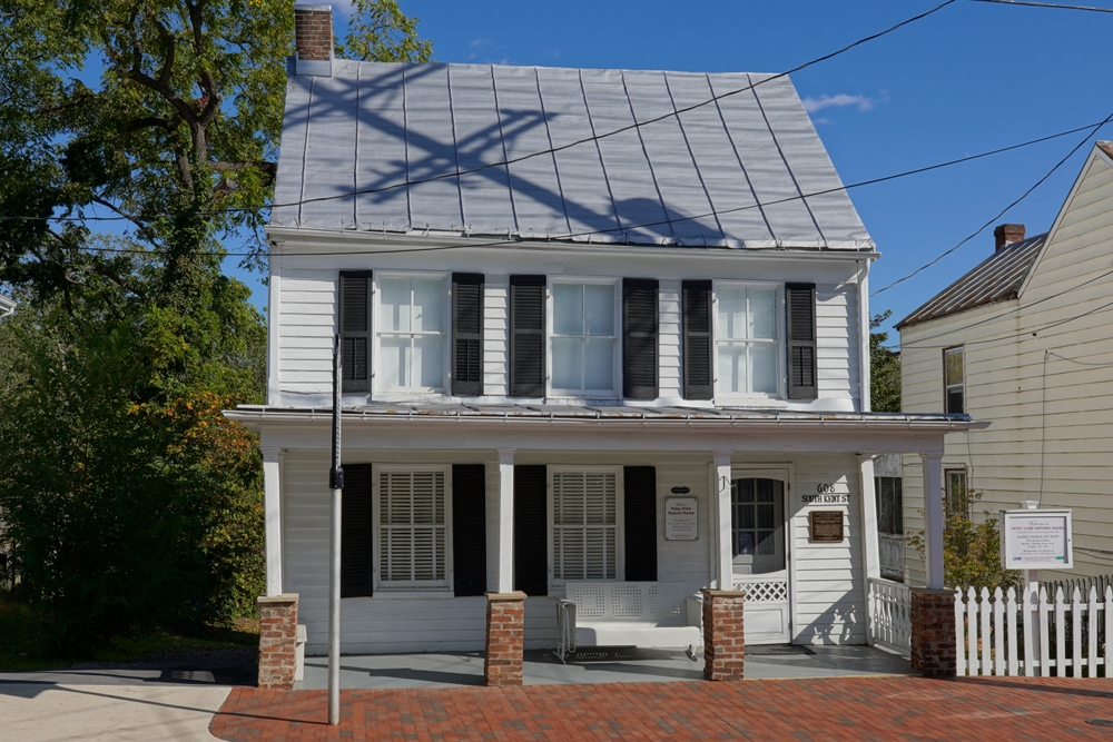 Historic charm meets Southern grace: A pristine white clapboard house with black shutters and a metal roof stands proudly on a sunlit street, its inviting porch and picket fence hinting at stories from a bygone era.