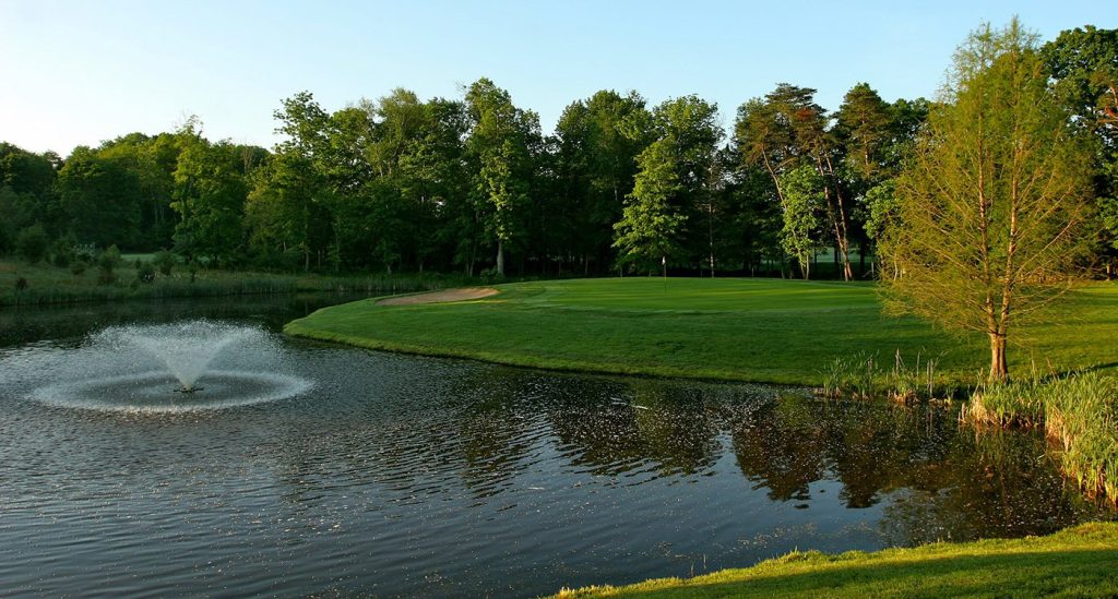 Serenity on the Green: A picturesque golf course hole bordered by a tranquil pond with a fountain, surrounded by lush trees on a calm summer day.