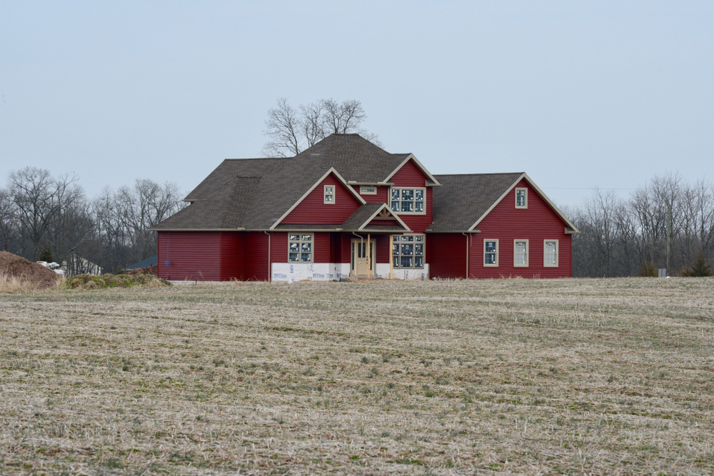 Charming red country house under construction amidst serene fields and tranquil trees.