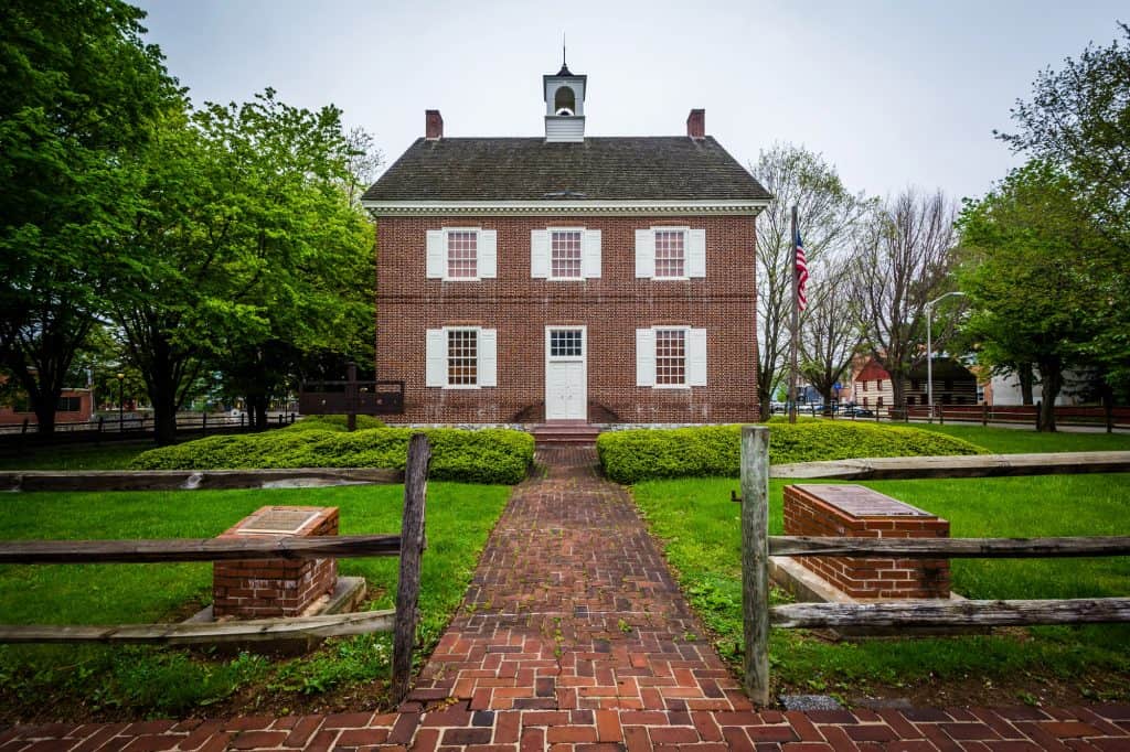 Historic Charm: Colonial-era brick courthouse stands proudly amidst manicured grounds, its white cupola and shutters a testament to early American architecture. A brick path and split-rail fence guide visitors to this well-preserved landmark, where the Stars and Stripes flutter in the breeze.