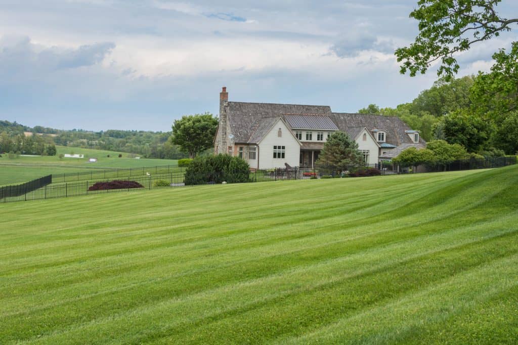 Elegant country estate with pristine striped lawn and wrought-iron fencing, featuring a cream-colored farmhouse with dormer windows and stone chimney, set against rolling hills and summer skies.
