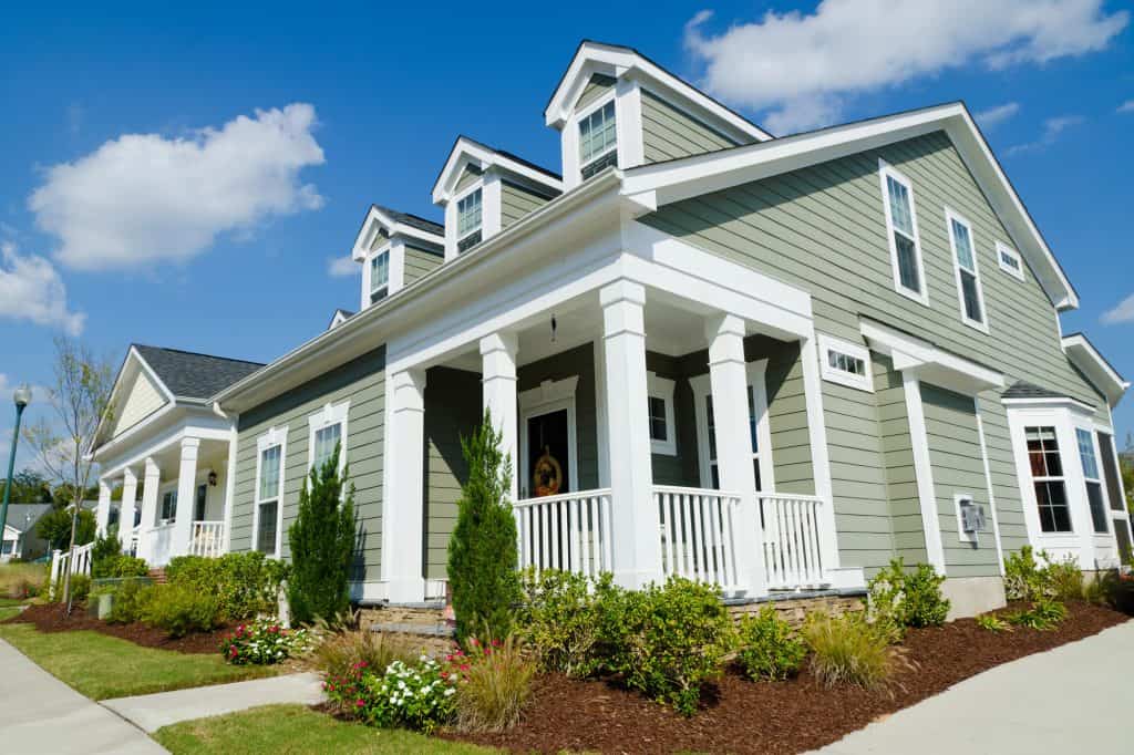 Classic American craftsman-style home featuring sage green siding, white columns, and dormer windows, with a welcoming front porch and meticulously landscaped garden beds dotted with flowering shrubs and ornamental grasses.