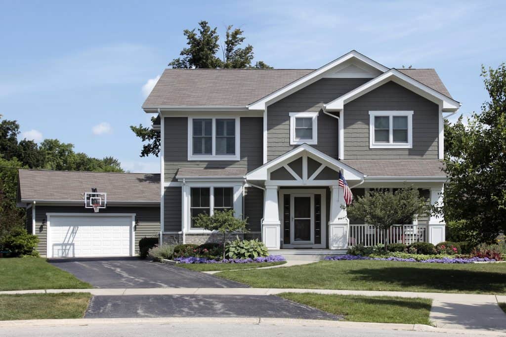 Contemporary two-story suburban home with charcoal gray siding, white trim, and a basketball hoop above the garage, featuring a welcoming front porch and colorful flower beds lining the entrance.