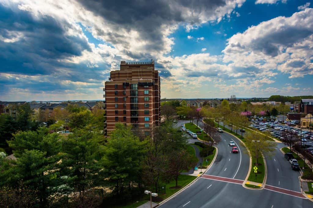 Modern high-rise apartment building stands tall against dramatic spring skies in a suburban landscape, with curving roads and parking lots framed by blooming trees and manicured greenery.