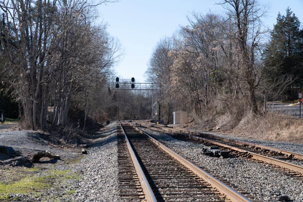 Railroad tracks stretch into the distance beneath a signal bridge, flanked by bare winter trees and gravel ballast on a clear day.